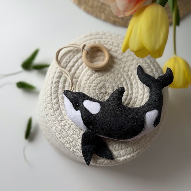 May include: A handmade felt orca whale toy in black and white, with a wooden ring and string for hanging. The toy is resting on a woven basket, with yellow tulips in the background.