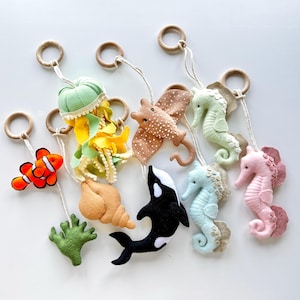 Puede incluir: Adornos de fieltro de criaturas marinas, incluyendo un pez payaso, una medusa, una raya, caballitos de mar y una orca. Cada adorno está unido a un anillo de madera con una cuerda blanca. Los adornos son de varios colores, como naranja, verde y rosa.