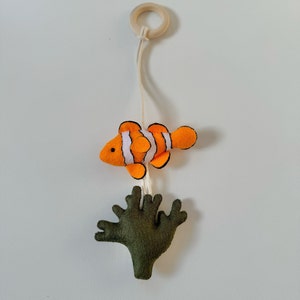 May include: A felt mobile featuring a clownfish with orange, white, and black stripes, and a green coral. The fish is suspended by a string from a wooden ring. The mobile is designed for a nursery or children's room.
