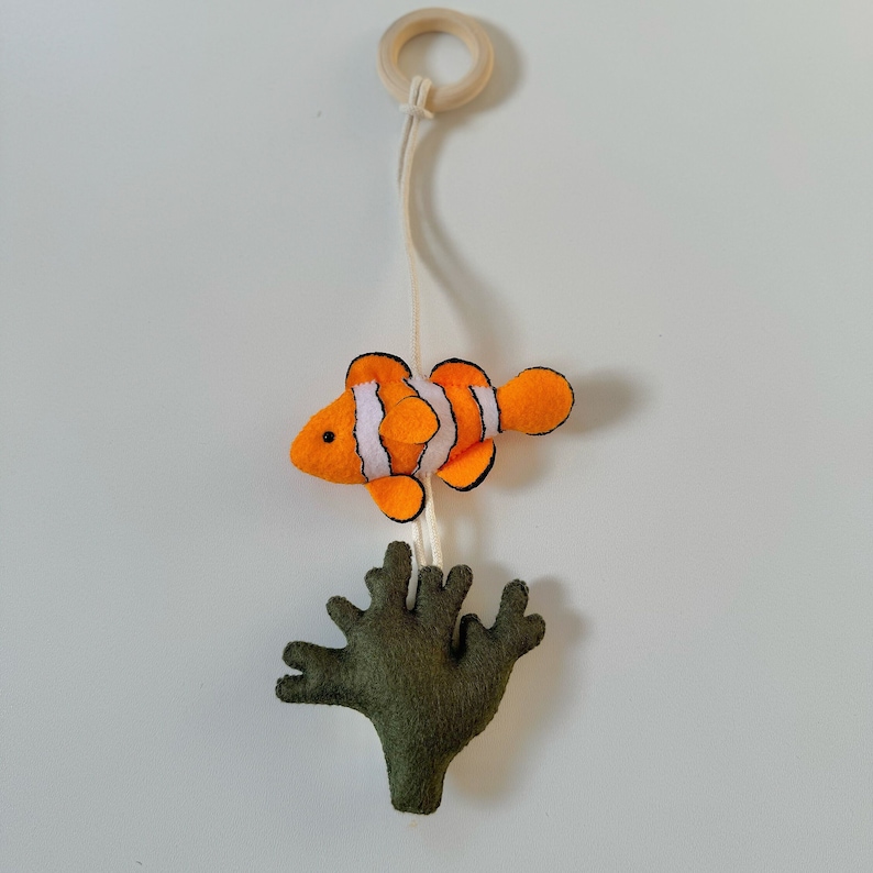 May include: A felt mobile featuring a clownfish with orange, white, and black stripes, and a green coral. The fish is suspended by a string from a wooden ring. The mobile is designed for a nursery or children's room.