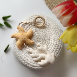 May include: A beige starfish and white coral-shaped felt ornaments hang from a wooden ring. The ornaments rest on a round, woven basket. The starfish is light yellow with small, dark dots. The coral is white.