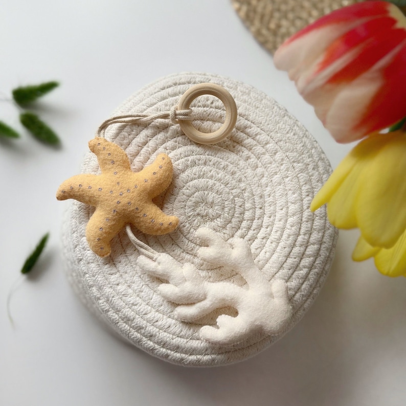 May include: A beige starfish and white coral-shaped felt ornaments hang from a wooden ring. The ornaments rest on a round, woven basket. The starfish is light yellow with small, dark dots. The coral is white.