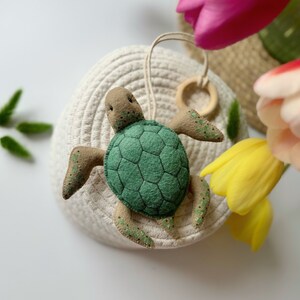 May include: A handmade felt turtle ornament with a green shell and tan head and flippers. The turtle has a wooden ring and a beige string for hanging. The ornament is placed on a white woven basket, with colorful tulips in the background.