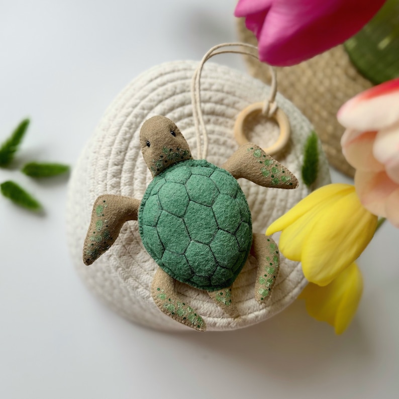 May include: A handmade felt turtle ornament with a green shell and tan head and flippers. The turtle has a wooden ring and a beige string for hanging. The ornament is placed on a white woven basket, with colorful tulips in the background.