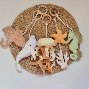 May include: A felt sea life mobile featuring a stingray, whale, jellyfish, starfish, seahorse, and coral. The sea creatures are in shades of beige, white, orange, and green, suspended from wooden rings and a woven circular background.