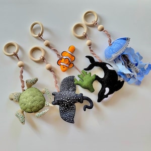 Könnte beinhalten: Handgefertigtes Baby-Mobile mit Meeresmotiven. Es zeigt eine Meeresschildkröte, einen Stechrochen, einen Clownfisch, einen Orca, grüne Algen und eine blaue Qualle, die an Holzringen hängen.