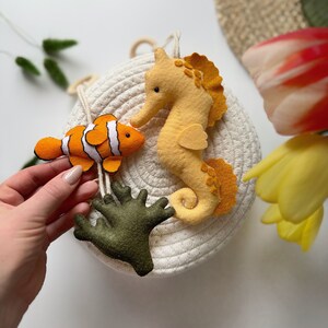 May include: A felt sea life mobile featuring a clownfish with orange and white stripes, a yellow seahorse, and green coral. The mobile is attached to a white rope basket, creating a nautical theme.