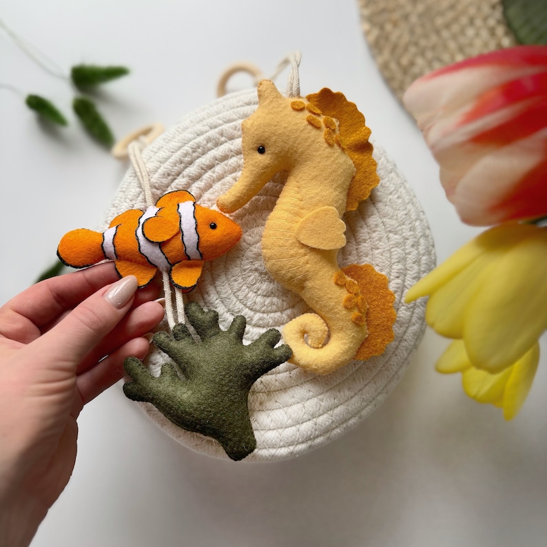 May include: A felt sea life mobile featuring a clownfish with orange and white stripes, a yellow seahorse, and green coral. The mobile is attached to a white rope basket, creating a nautical theme.