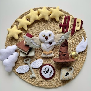 May include: A felt craft featuring Harry Potter themed characters and objects.  The craft includes a white owl, a sorting hat, a golden snitch, a book, a key, a cloud, a 9 3/4 platform sign, and stars.  The craft is displayed on a woven straw mat.
