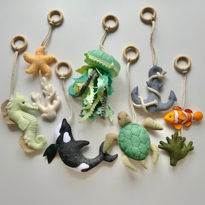 May include: A collection of felt sea creatures, including a seahorse, starfish, jellyfish, orca, turtle, clownfish, anchor, and coral, each suspended by a wooden ring and string. The creatures are in various colors, such as green, orange, and gray.