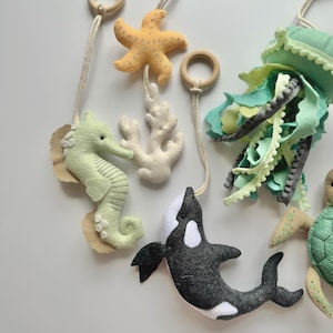 May include: A felt sea-themed mobile featuring a seahorse, starfish, coral, jellyfish, and orca. The mobile includes wooden rings and is in shades of green, yellow, white, and black. The seahorse is approximately 4 inches tall.