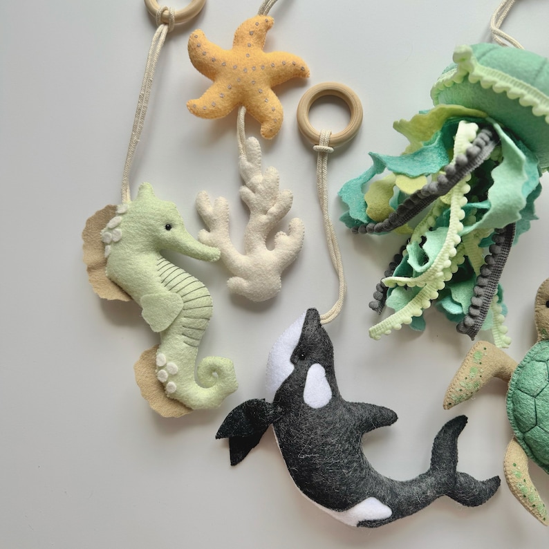 May include: A felt sea-themed mobile featuring a seahorse, starfish, coral, jellyfish, and orca. The mobile includes wooden rings and is in shades of green, yellow, white, and black. The seahorse is approximately 4 inches tall.