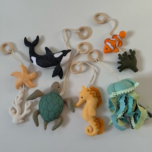 May include: A collection of felt sea creatures, including a black and white orca, a sea turtle, a seahorse, a starfish, a clownfish, a jellyfish, and coral. Each creature is suspended by a string and wooden ring, creating a mobile.