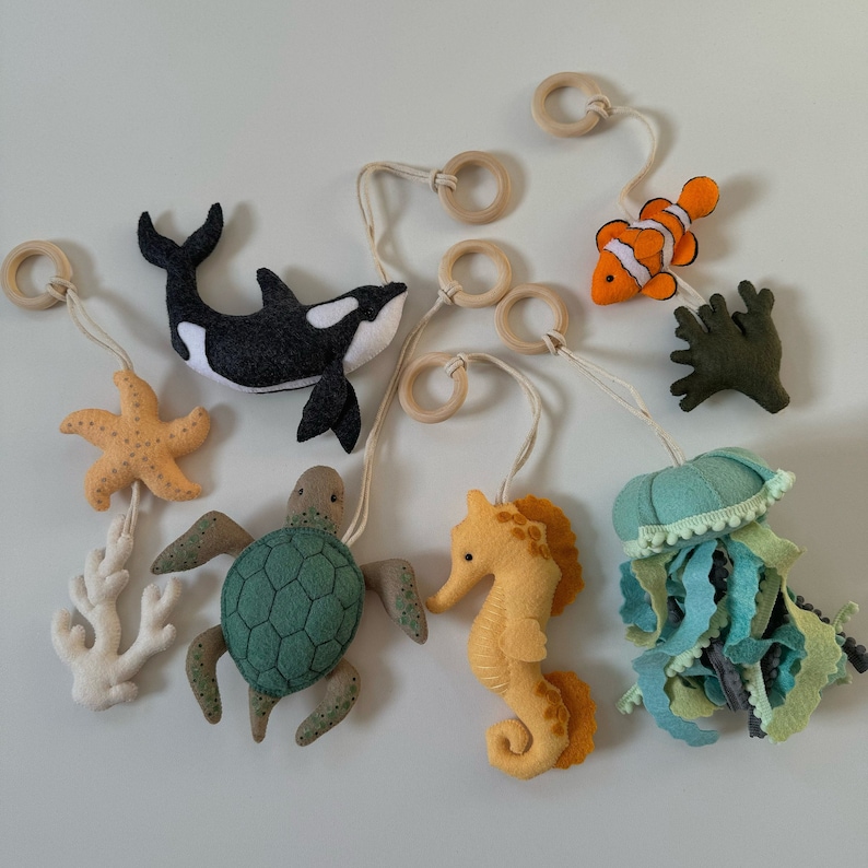 May include: A collection of felt sea creatures, including a black and white orca, a sea turtle, a seahorse, a starfish, a clownfish, a jellyfish, and coral. Each creature is suspended by a string and wooden ring, creating a mobile.