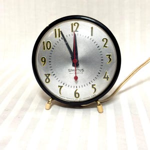 Könnte beinhalten: Eine Vintage Spartus Uhr mit Selbstaufzug, silbernem Zifferblatt, schwarzem Rahmen und goldfarbenen Beinen. Die Uhr hat goldene Ziffern und Zeiger, mit einem roten Sekundenzeiger. Die Uhrzeit ist ungefähr 12:00 Uhr.