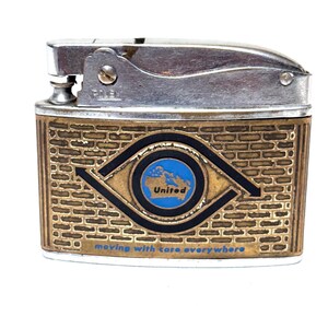 May include: Vintage metal lighter with a silver top and a gold-toned body featuring a brick pattern. The design includes a blue and gold emblem with the word "United" and the phrase "moving with care everywhere."