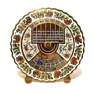 May include: A round brass 28-year calendar with a scalloped edge and white enamel floral patterns in red, blue, and green. The center features a grid of days and dates, with a rotating mechanism below displaying years and months in blue and red text.
