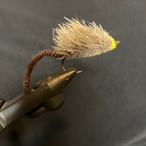 May include: A brown and gray fly fishing lure with a yellow head and a brown tail. The lure is tied to a fishing hook and is attached to a black fishing rod.