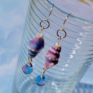 May include: A pair of seashell earrings with a gradient of purple and blue hues. The earrings have gold-colored hooks and small, round, blue glass beads dangling below the shells. The earrings are set against a blurred background.