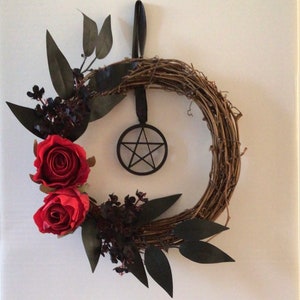 May include: A circular grapevine wreath featuring two red roses, black leaves, and dark berries. A black pentagram is suspended from a black ribbon. Ideal for door or wall decoration.