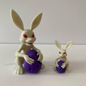 May include: Two off-white rabbit figurines with red eyes, each clutching a purple egg with a carved face. The larger rabbit is approximately 10 cm tall, while the smaller one is about 5 cm tall. The figurines are set against a plain white background.