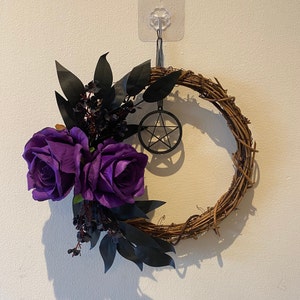 May include: A circular grapevine wreath featuring two large purple roses, black leaves, and dark berries. A black metal pentagram charm is suspended from the wreath. The wreath is displayed on a white wall.