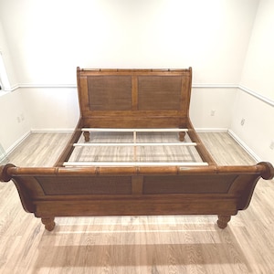 May include: A wooden sleigh bed frame with a medium brown finish and a faux bamboo texture on the frame edges. The headboard and footboard feature woven cane panels. The bed includes a set of light-colored wooden slats for mattress support.