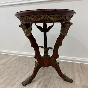 May include: A round wooden side table featuring a dark brown finish and gold-toned metal accents. The table has three legs adorned with eagle-shaped carvings at the top and claw-style feet at the base, with a central eagle figure connecting the legs.