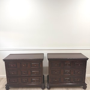 May include: Two matching dark brown wooden chests of drawers. Each chest has nine drawers with small, round knobs. The chests have ornate carvings along the base and top edges, and sit on decorative feet.