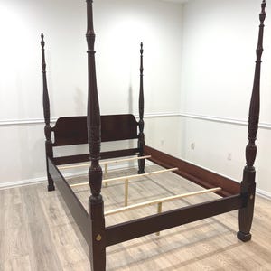 May include: A dark brown four-poster bed frame with a headboard. The bed frame has four tall, ornate posts with decorative finials. The bed frame is made of wood and is set on a light-colored wooden floor.