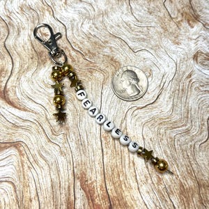 May include: A silver-tone keychain with a beaded charm spelling "FEARLESS" in white and black letter beads. The charm includes gold-colored beads and star-shaped accents. A quarter is next to the keychain.