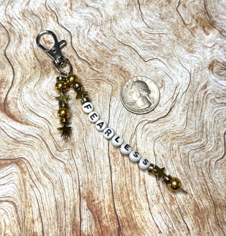 May include: A silver-tone keychain with a beaded charm spelling "FEARLESS" in white and black letter beads. The charm includes gold-colored beads and star-shaped accents. A quarter is next to the keychain.