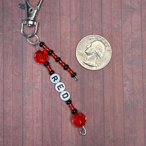 以下が含まれることがあります： 白い文字ビーズで「RED」と書かれたビーズキーホルダー。 赤と黒のビーズ、シルバーの留め金、シルバーのジャンプリングが付いています。 キーホルダーの隣にはコインがあります。