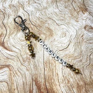 May include: A keychain with a silver clasp and a beaded strand. The strand features gold-colored beads and star-shaped charms, along with white letter beads spelling the word "FEARLESS". The background is a wooden surface.