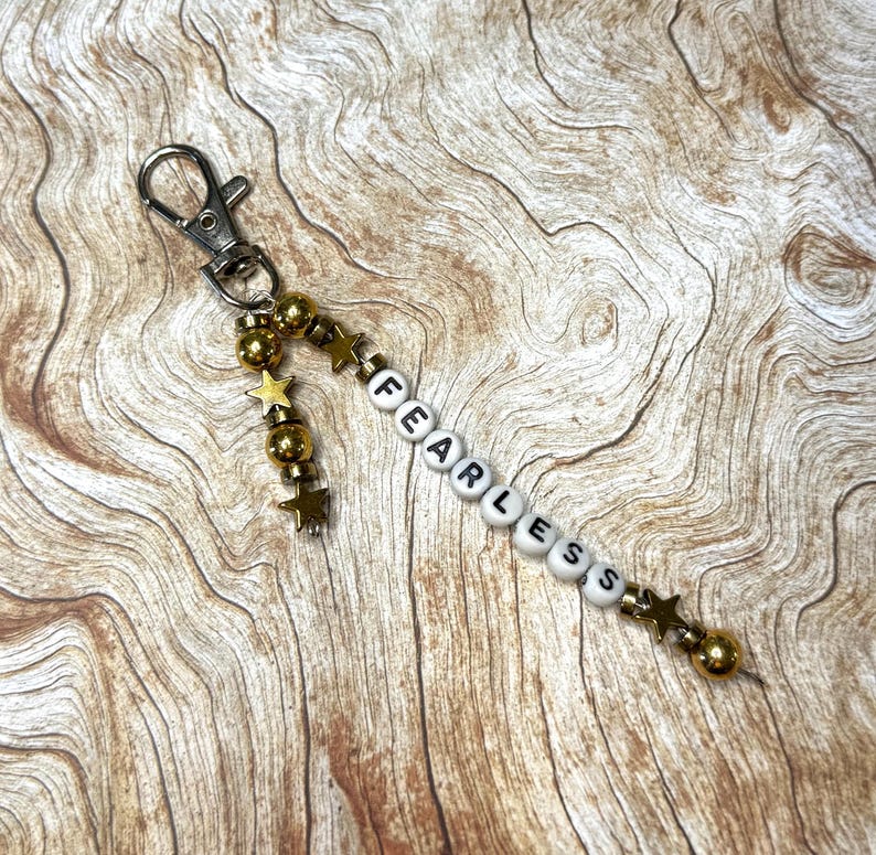 May include: A keychain with a silver clasp and a beaded strand. The strand features gold-colored beads and star-shaped charms, along with white letter beads spelling the word "FEARLESS". The background is a wooden surface.
