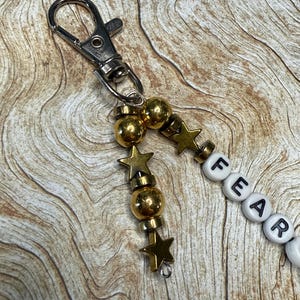 May include: A keychain with a silver clasp, gold beads, star charms, and white letter beads spelling "FEAR". The beads and charms are strung together, creating a decorative accessory.