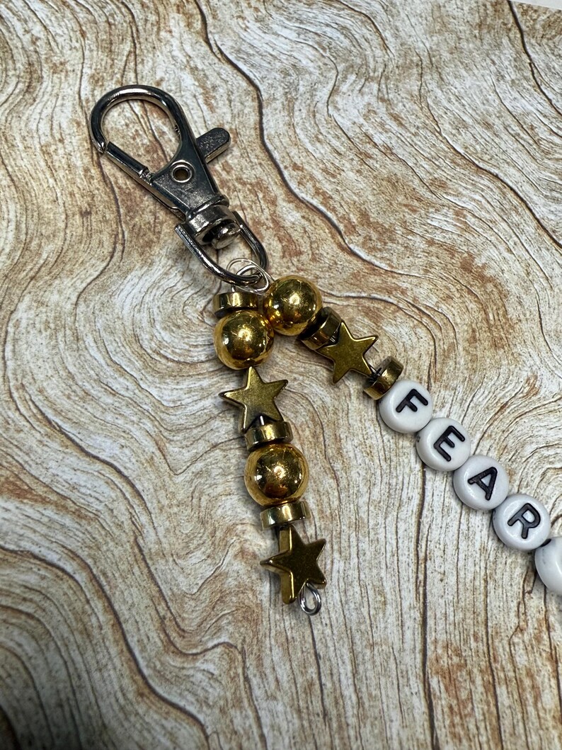 May include: A keychain with a silver clasp, gold beads, star charms, and white letter beads spelling "FEAR". The beads and charms are strung together, creating a decorative accessory.