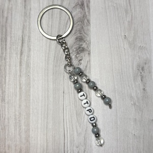 May include: A silver keychain with a chain and beaded accents. The beads are gray and clear, with white letter beads spelling "TTPD". The keychain is on a light wood grain background.