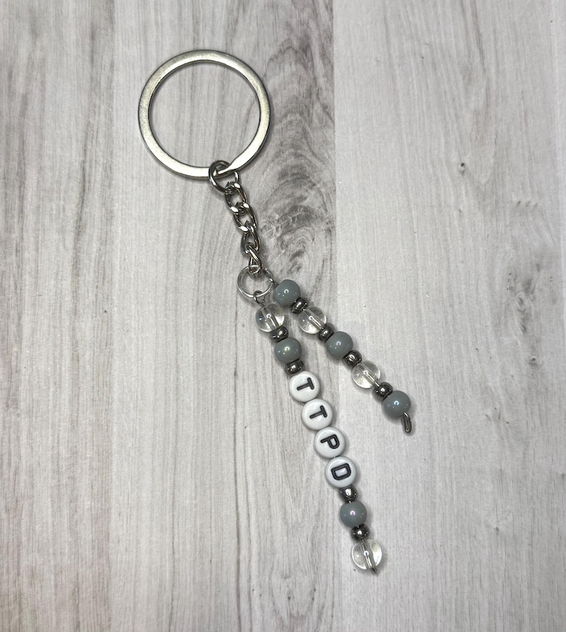 May include: A silver keychain with a chain and beaded accents. The beads are gray and clear, with white letter beads spelling "TTPD". The keychain is on a light wood grain background.