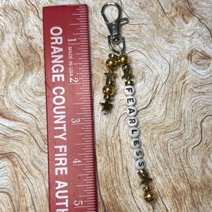 May include: A silver keychain with a lobster clasp, gold beads, and star charms. The keychain has white letter beads that spell "FEARLESS". A red ruler with the text "ORANGE COUNTY FIRE AUTH" is in the image.