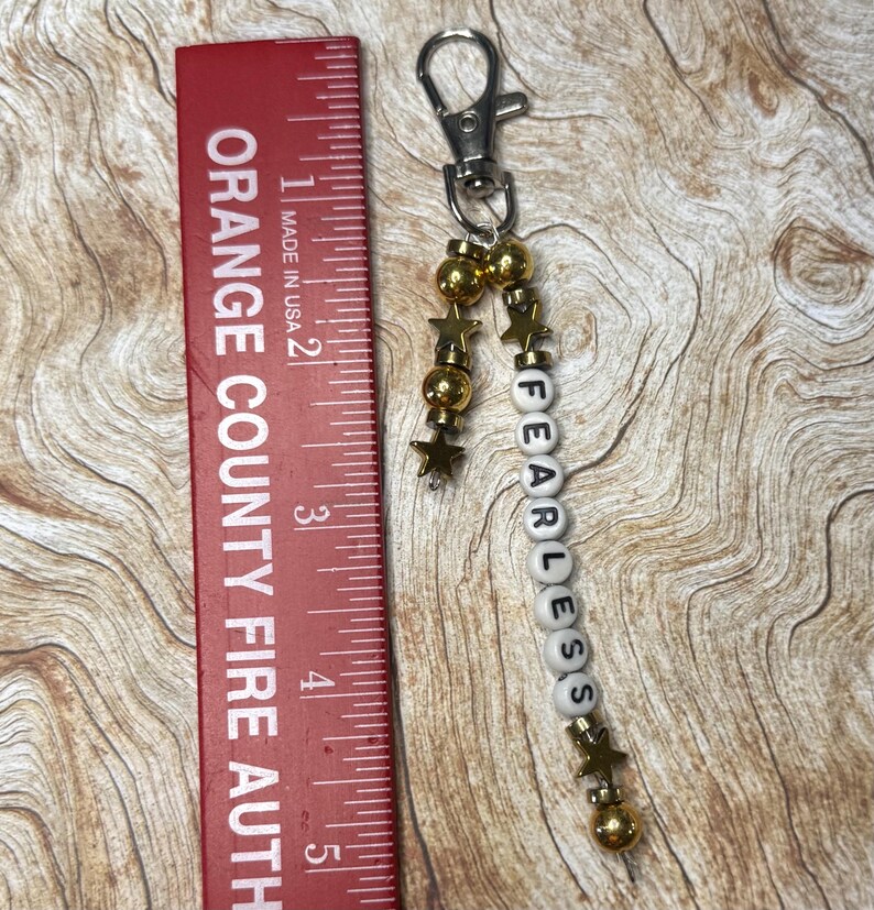 May include: A silver keychain with a lobster clasp, gold beads, and star charms. The keychain has white letter beads that spell "FEARLESS". A red ruler with the text "ORANGE COUNTY FIRE AUTH" is in the image.