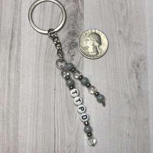 May include: A silver keychain with a chain and beaded accents. The keychain has gray and clear beads, with the letters "TTPD" spelled out on white letter beads. A quarter is next to the keychain.