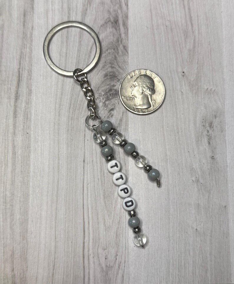 May include: A silver keychain with a chain and beaded accents. The keychain has gray and clear beads, with the letters "TTPD" spelled out on white letter beads. A quarter is next to the keychain.