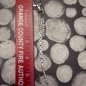Op de afbeelding: Een zilverkleurige sleutelhanger met een kralenketting en het woord "FOLKLORE" gespeld in witte letterkralen. De sleutelhanger ligt naast een rode liniaal en is ongeveer 13 cm lang. De liniaal leest "ORANGE COUNTY FIRE AUTHOR".