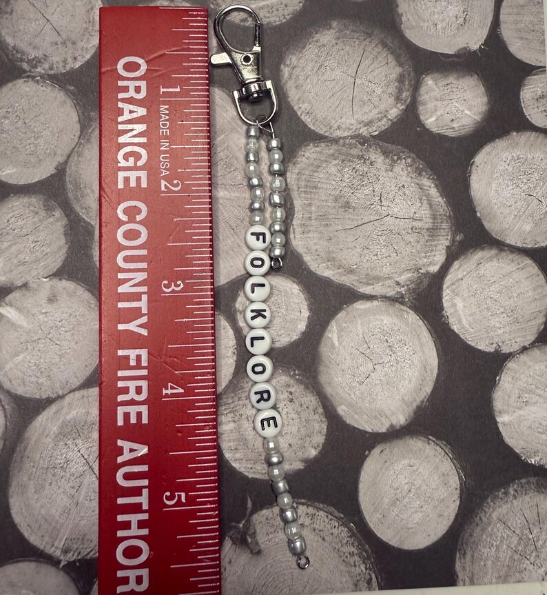 Op de afbeelding: Een zilverkleurige sleutelhanger met een kralenketting en het woord "FOLKLORE" gespeld in witte letterkralen. De sleutelhanger ligt naast een rode liniaal en is ongeveer 13 cm lang. De liniaal leest "ORANGE COUNTY FIRE AUTHOR".