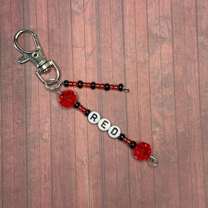 以下が含まれることがあります： 白い文字ビーズで「RED」と書かれたビーズのキーホルダー。キーホルダーは赤と黒のビーズで、シルバーの留め金が付いています。背景は木製の表面です。