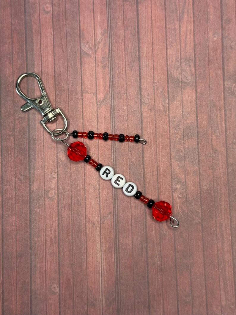 以下が含まれることがあります： 白い文字ビーズで「RED」と書かれたビーズのキーホルダー。キーホルダーは赤と黒のビーズで、シルバーの留め金が付いています。背景は木製の表面です。