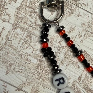 Op de afbeelding: Een kralen lanyard met een zilveren sluiting. De lanyard heeft zwarte en rode kralen, met witte letterkralen die een woord spellen. De kralen zijn aan elkaar geregen, waardoor een decoratief en functioneel accessoire ontstaat.