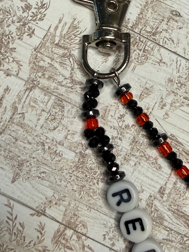Op de afbeelding: Een kralen lanyard met een zilveren sluiting. De lanyard heeft zwarte en rode kralen, met witte letterkralen die een woord spellen. De kralen zijn aan elkaar geregen, waardoor een decoratief en functioneel accessoire ontstaat.