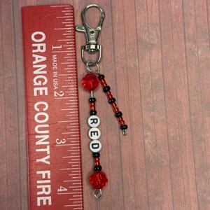 以下が含まれることがあります： 白と黒の文字ビーズで「RED」と書かれた赤いビーズのキーホルダー。キーホルダーには赤と黒のビーズ、シルバーの留め金、小さな赤い定規が含まれています。